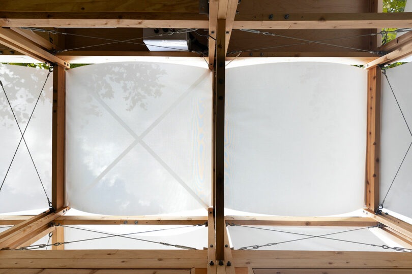 View looking up at a wooden pergola structure with white fabric panels stretched across the top, supported by metal cables.