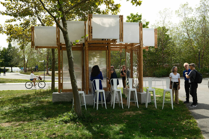 A small open-air wooden pavilion with white panels and chairs stands in a park, with several people gathered around and a cyclist passing by in the background.