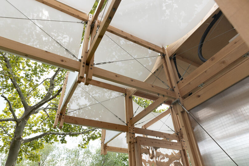 Close-up view of a wooden structure with metal cables and white canopy panels, set outdoors near leafy trees.