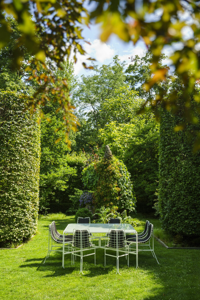 A metal table with six chairs is set on a green lawn, surrounded by tall, manicured hedges and lush garden foliage under a partly cloudy sky.