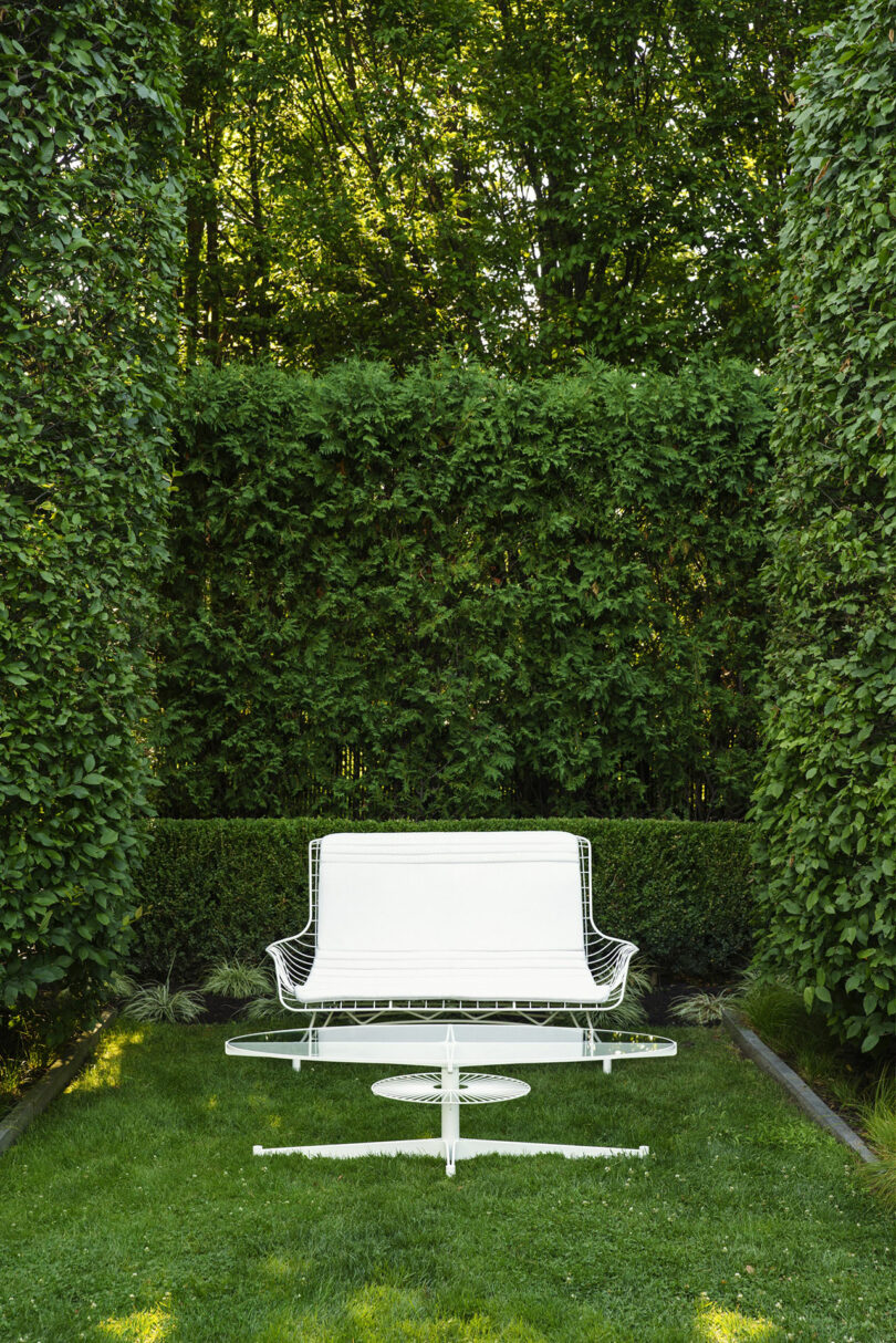 White metal outdoor bench and table set on green grass, surrounded by tall, dense hedges and trees under natural daylight.