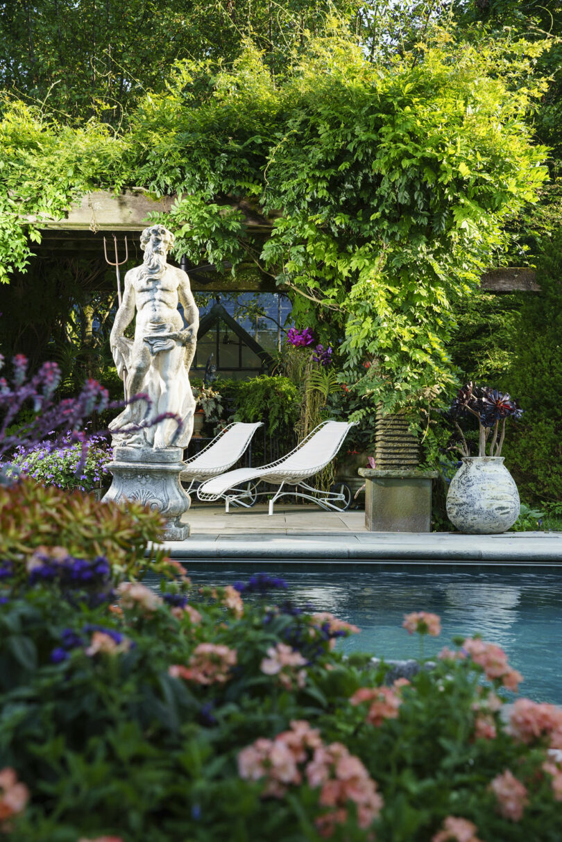 A garden scene with a pool in the foreground, a classical stone statue, two white lounge chairs, lush plants, and a pergola covered in greenery.