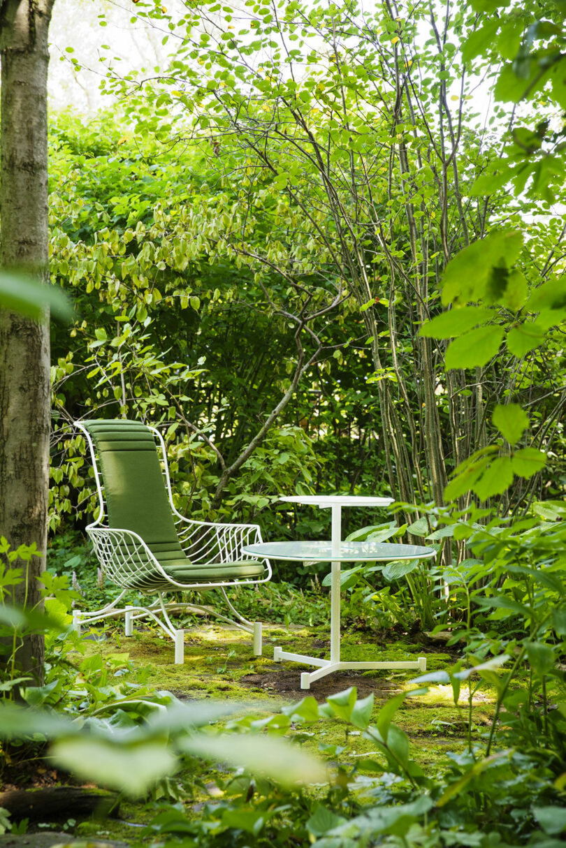 A green outdoor chair and a white table sit on a mossy patio surrounded by dense green foliage and trees in a garden.
