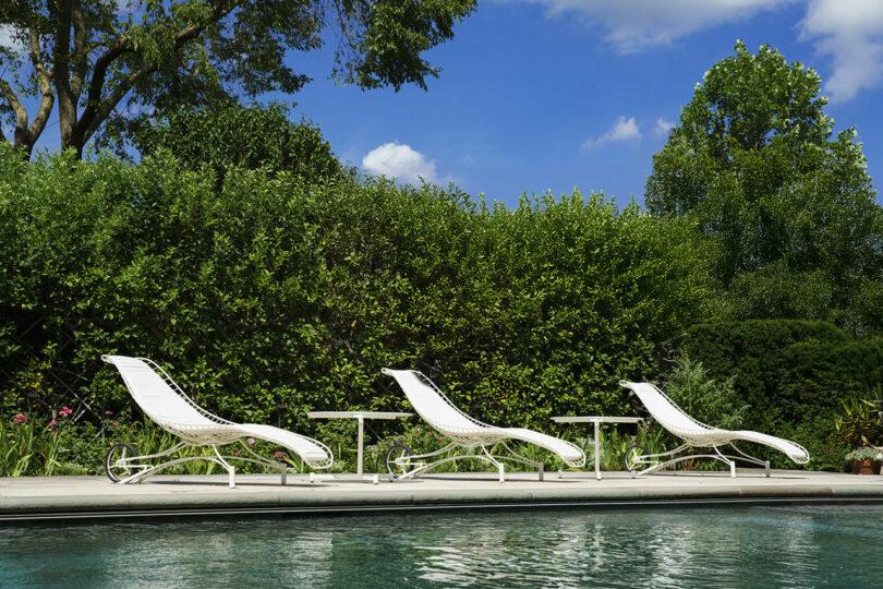 Three white lounge chairs and two small white tables are lined up beside a pool, with green bushes and trees in the background under a blue sky.
