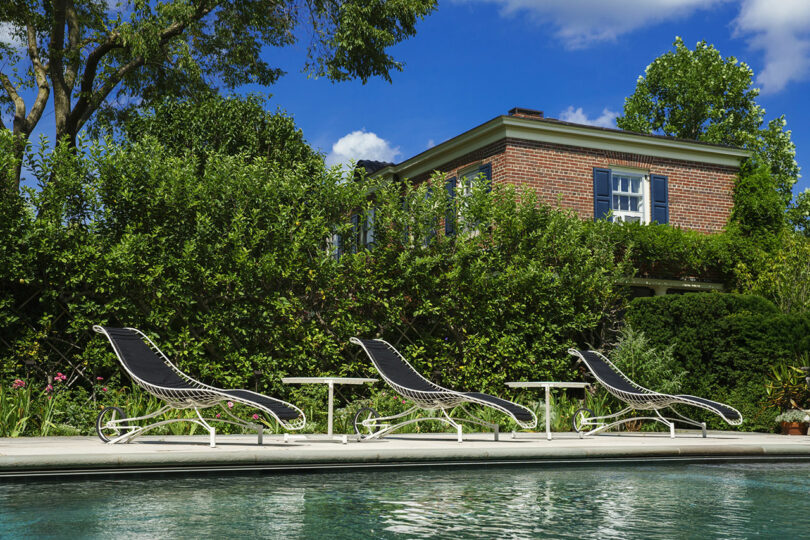 Three modern lounge chairs with side tables are arranged by a swimming pool, with a brick house and green bushes in the background under a blue sky.