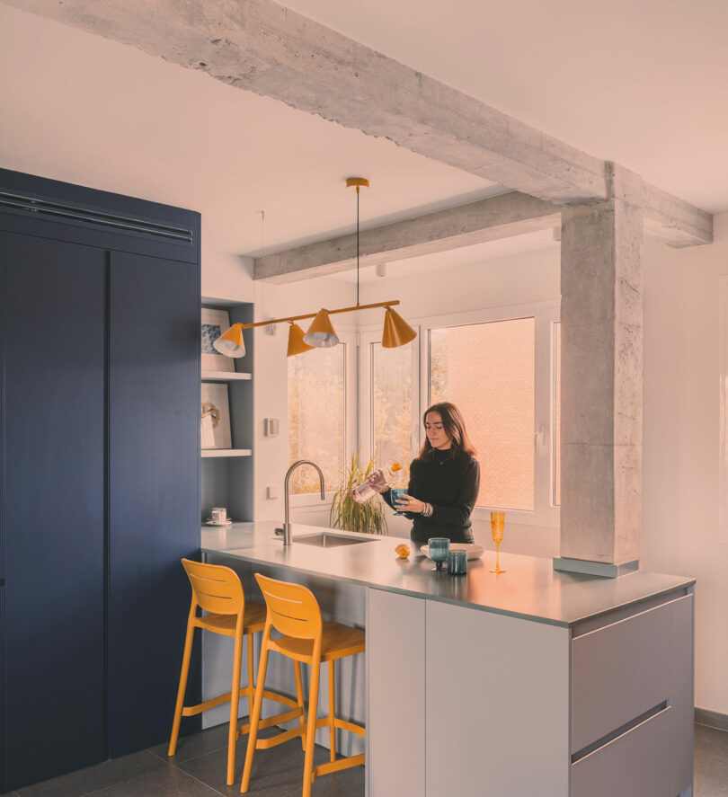 A woman stands in a modern kitchen with yellow chairs and pendant light, pouring a drink at the counter near a window.