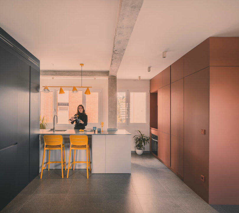 A person stands at a modern kitchen island with yellow stools, surrounded by dark and rust-colored cabinets, under pendant lights, with large windows in the background.