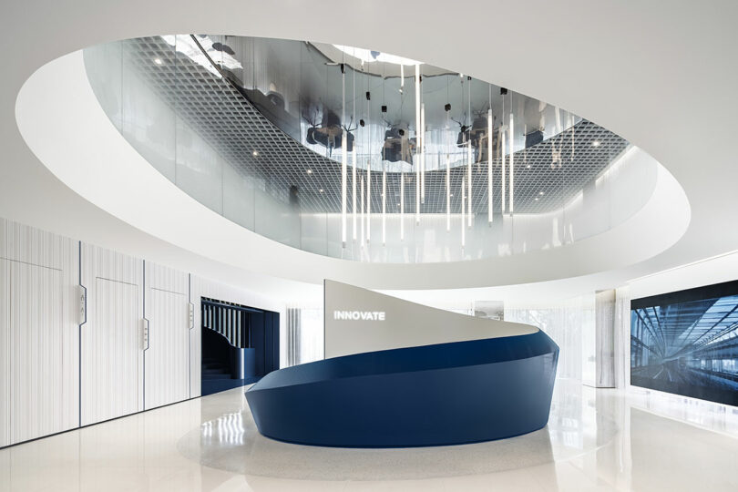 Modern office lobby with a blue-and-white curved reception desk, white walls, glossy floor, and ceiling lights reflected in a large oval opening. The word "INNOVATE" is illuminated on the wall.