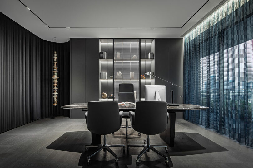 Modern office with a large desk, two guest chairs, a computer, shelves with decor, floor-to-ceiling windows, and dark minimalist decor.