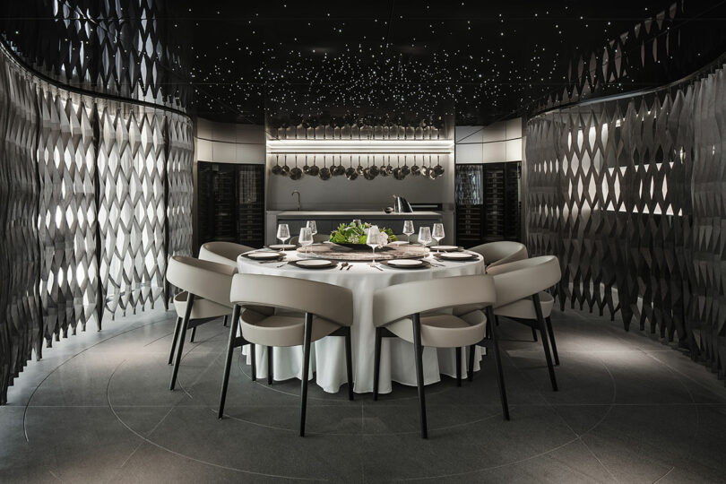 A round dining table set for six with modern white chairs sits in a dim, stylish room with dark, patterned walls and a ceiling featuring small, star-like lights.