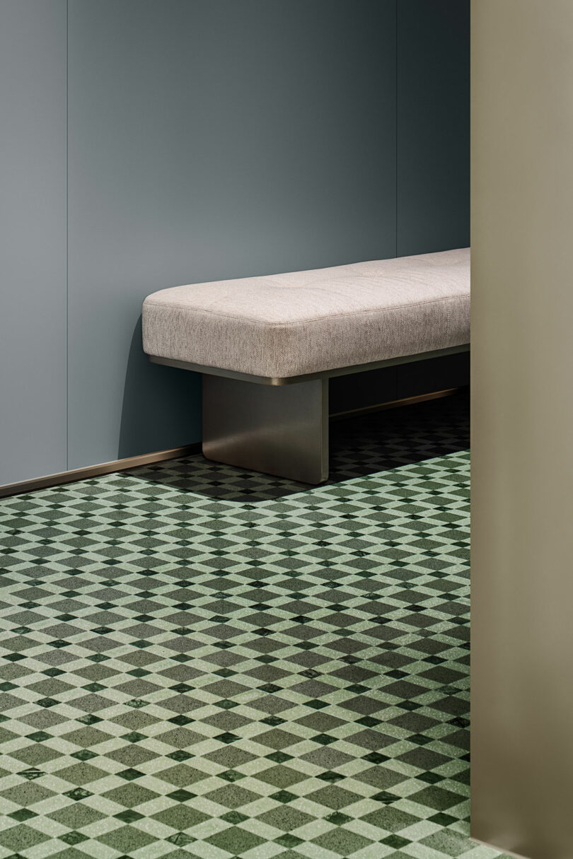 A minimalist bench with a light cushion sits against a gray wall, with a green geometric patterned floor and a partial beige partition in view.