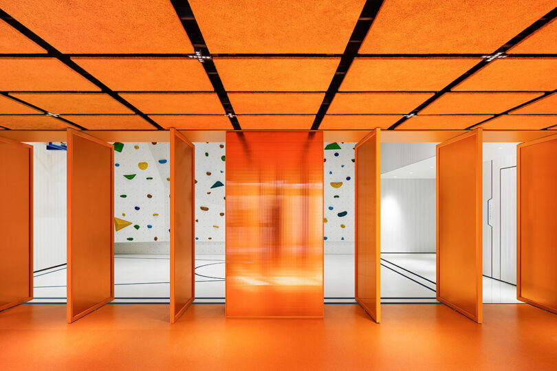 A brightly lit interior with orange panels and ceiling, leading to a gym space with a white wall featuring colorful climbing holds.