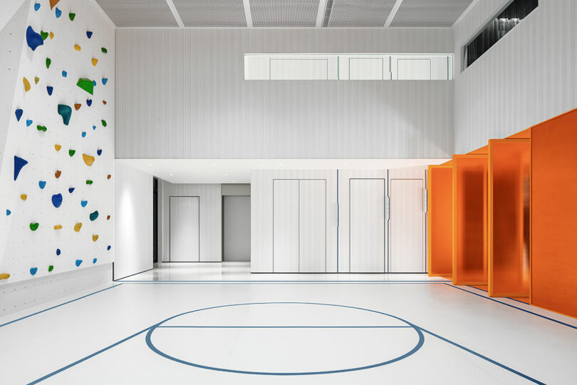 Modern indoor gym with white walls, a blue-lined basketball court, a colorful climbing wall, and a row of orange cubicles on the right.