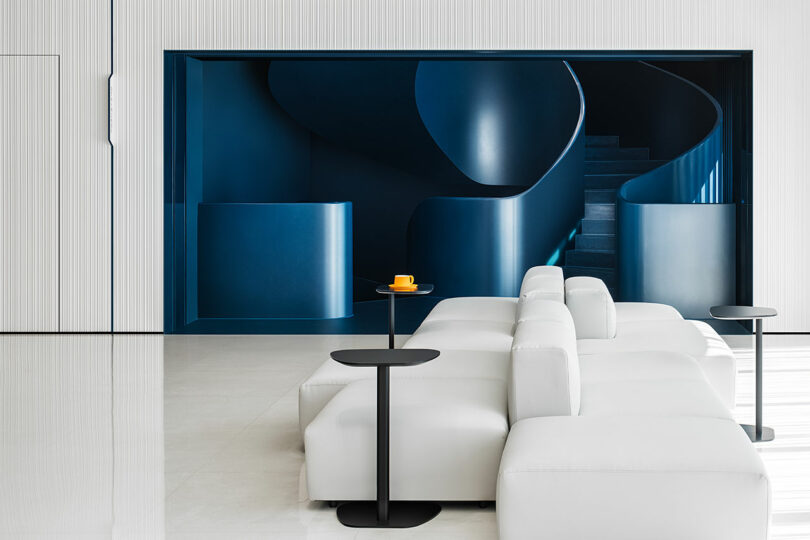 A modern interior with white modular seating and black tables in front of a deep blue, sculptural spiral staircase. A yellow cup and saucer sit on one table.
