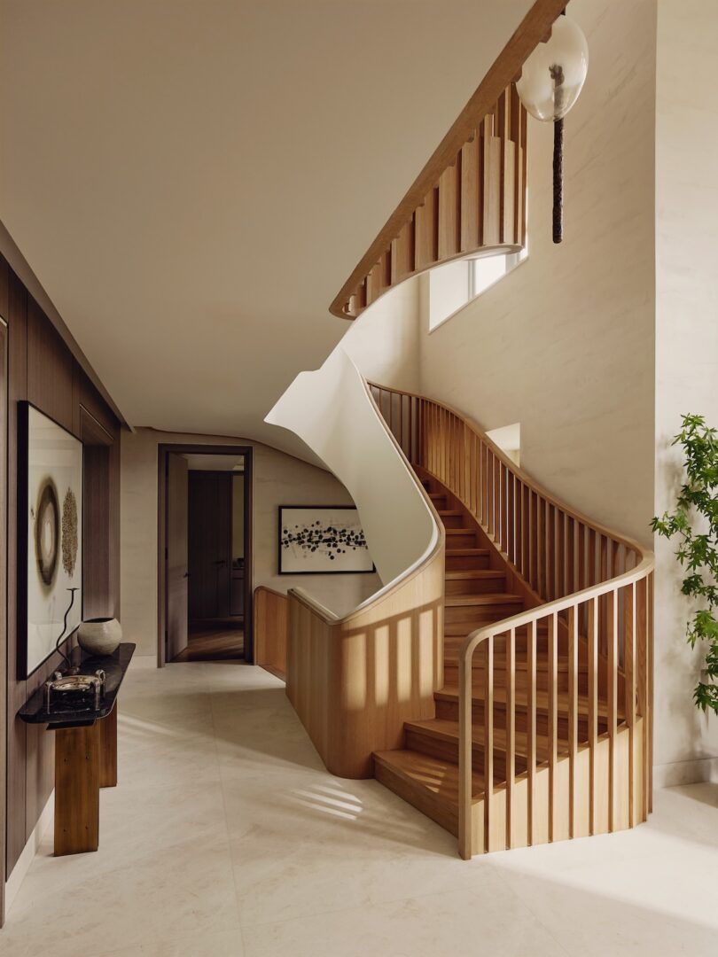 Curved wooden staircase with vertical railings in a modern, well-lit interior featuring beige floors, wall art, and a potted plant.