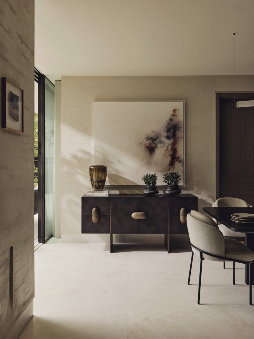 Modern dining area with a dark sideboard, two potted plants, a brown glass vase, and abstract wall art. Natural light enters from a large window, illuminating the neutral-toned room.