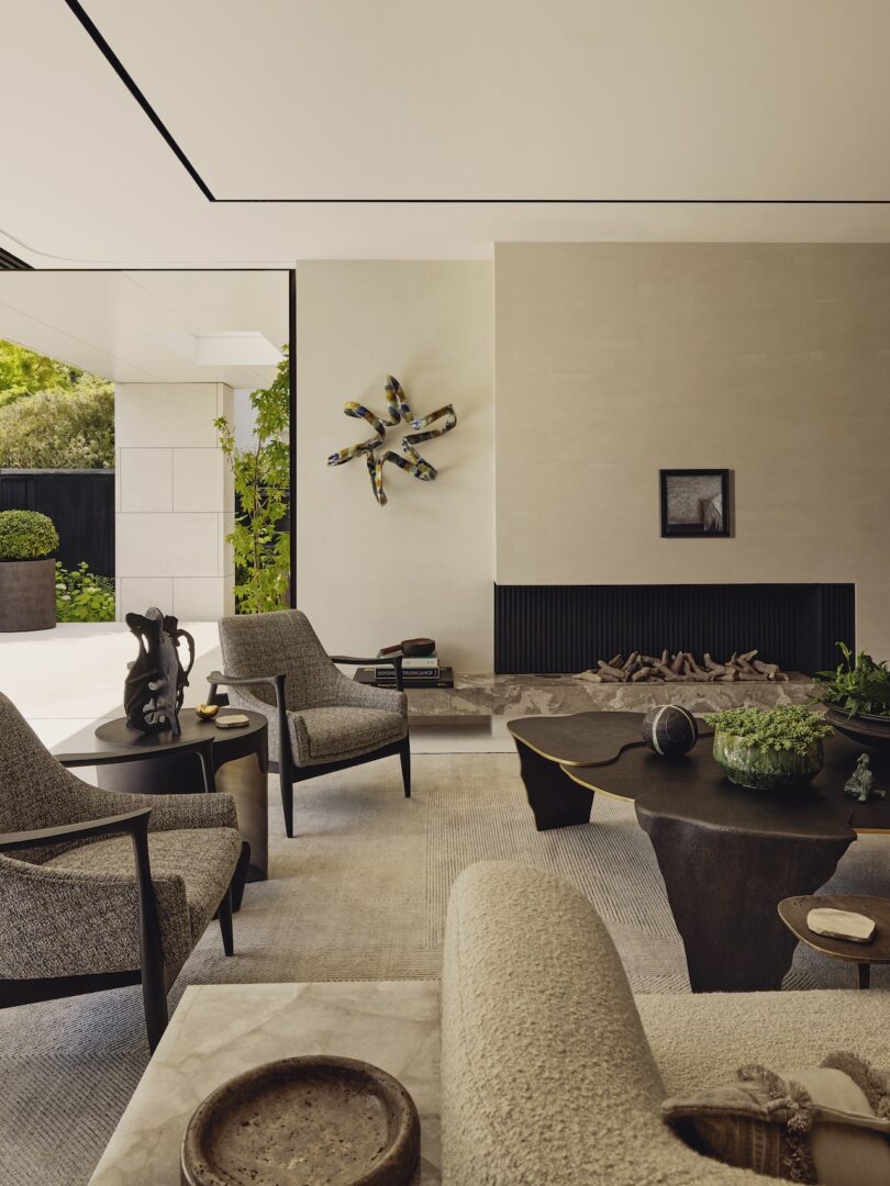 Modern living room with neutral tones, contemporary furniture, a wall-mounted abstract sculpture, framed art, and a fireplace with stacked wood, opening to a green outdoor area.