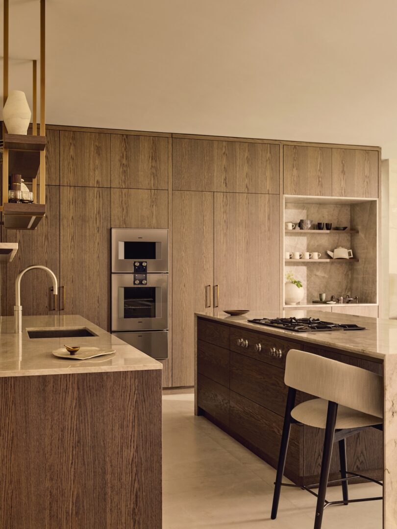 Modern kitchen with wood cabinetry, built-in stainless steel appliances, a central island with a cooktop and seating, and open shelves displaying cups and bowls.