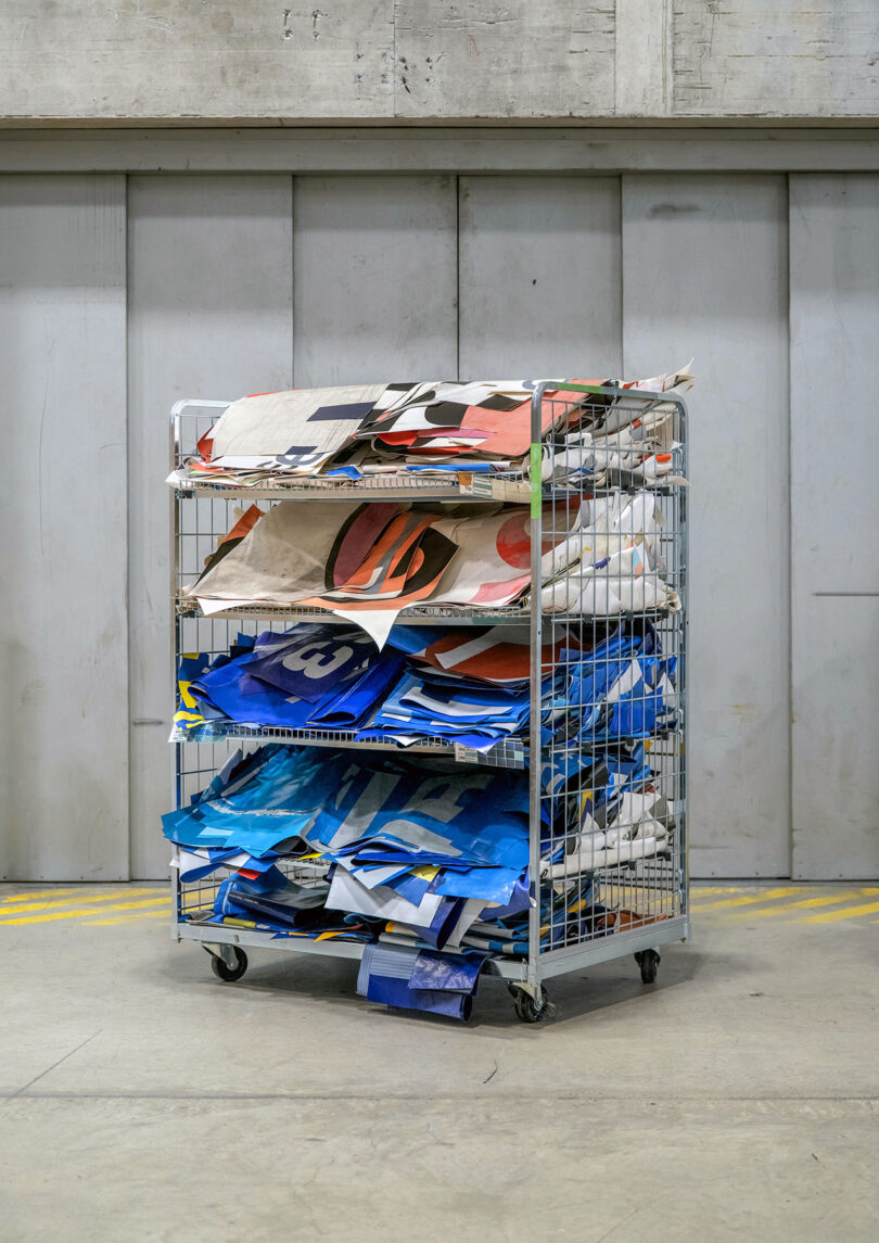 A metal cart with three shelves holds stacked pieces of folded, colorful printed fabric in an industrial setting.