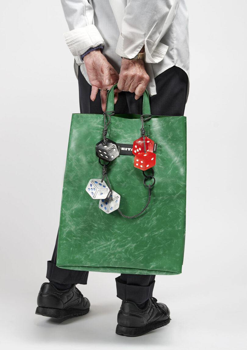 A person stands holding a green textured tote bag decorated with large red, black, and white dice keychains. The person wears black shoes, dark pants, and a white shirt.