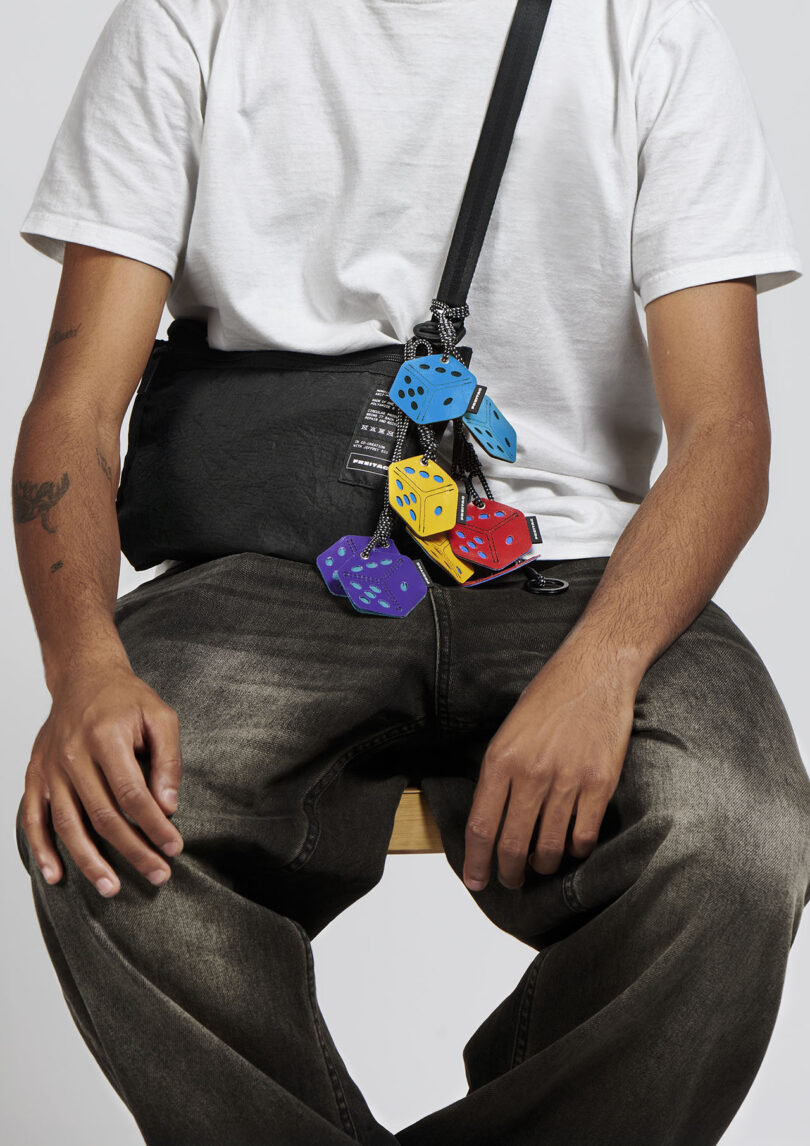 Person wearing a white t-shirt and dark jeans sits on a stool with a black crossbody bag decorated with several large, colorful plush dice hanging from its strap.