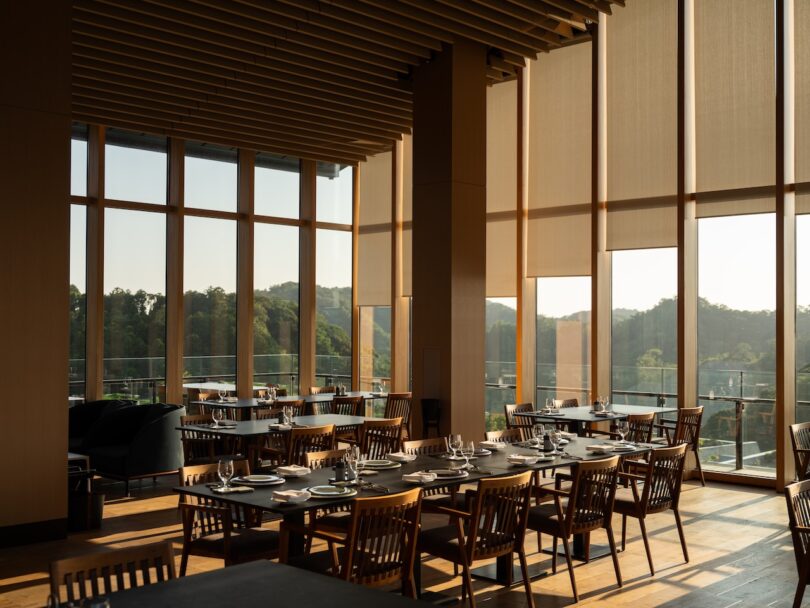 A modern restaurant dining area with wooden tables and chairs, set for service, large floor-to-ceiling windows, and natural light streaming in.
