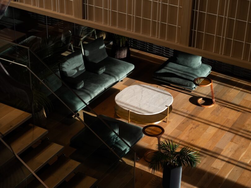 Modern living room with green velvet chairs, a white marble coffee table, wooden side tables, and potted plants on a wood floor, lit by natural sunlight.