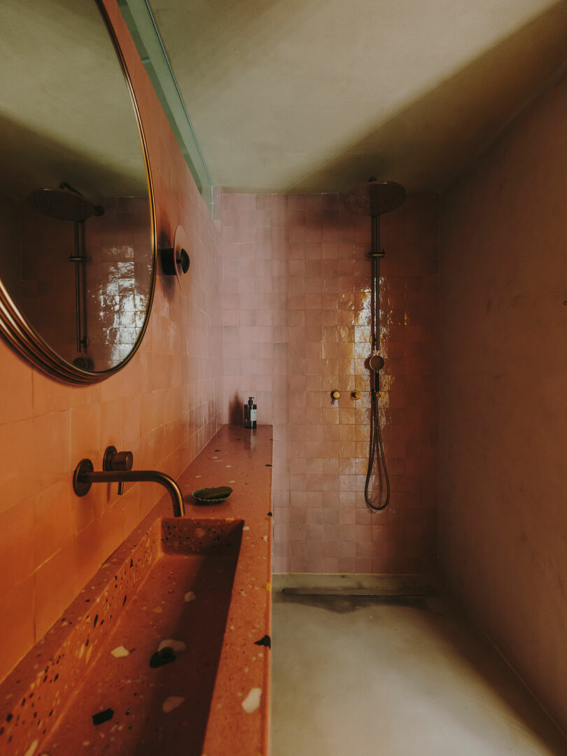 Modern bathroom with pink tiled wall, large round mirror, terrazzo countertop, wall-mounted faucet, and a walk-in shower with a rain shower head.