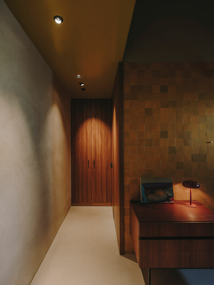 A hallway with beige walls and ceiling spotlights leads to wooden doors; a wooden desk with a red lamp sits on the right against a tiled wall.