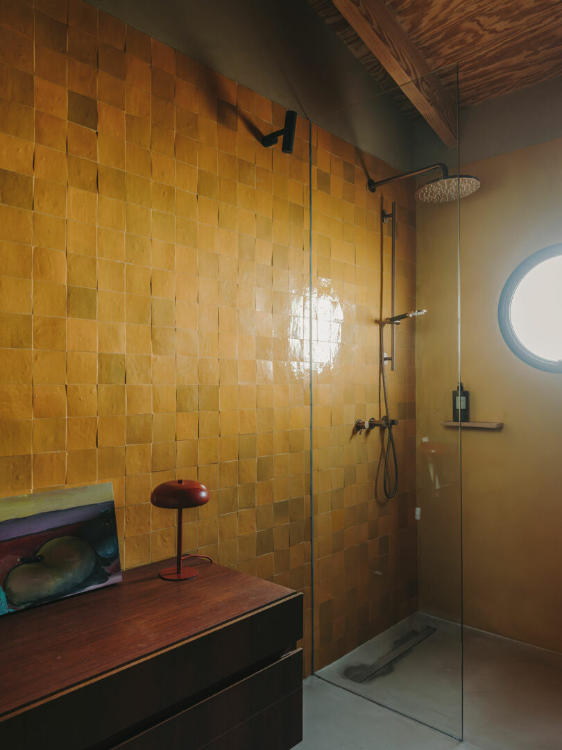 Modern bathroom with yellow-tiled wall, glass shower enclosure, circular window, wooden vanity, and red table lamp.