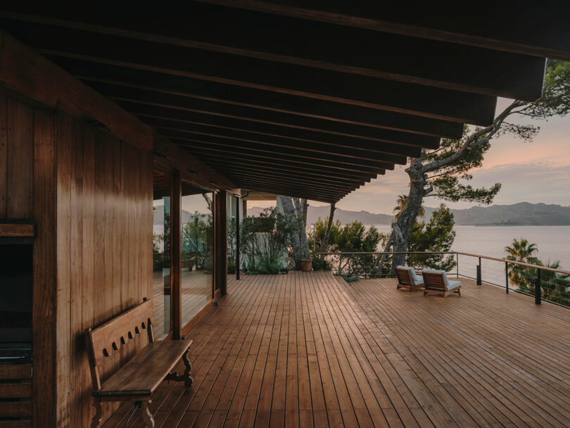 A wooden terrace with a bench and two lounge chairs overlooks a body of water and distant hills, surrounded by trees at sunset.