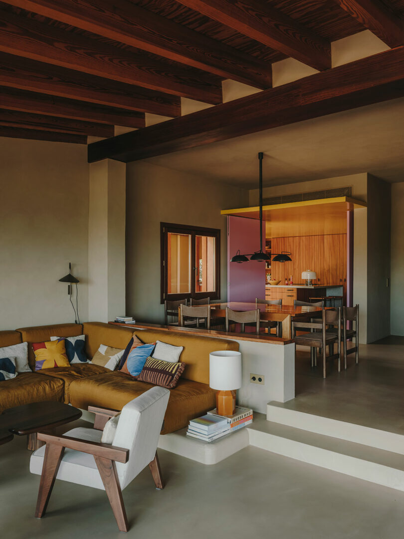 A modern living and dining area with a mustard sectional sofa, wooden furniture, exposed beam ceiling, and a kitchen in the background with colorful walls.