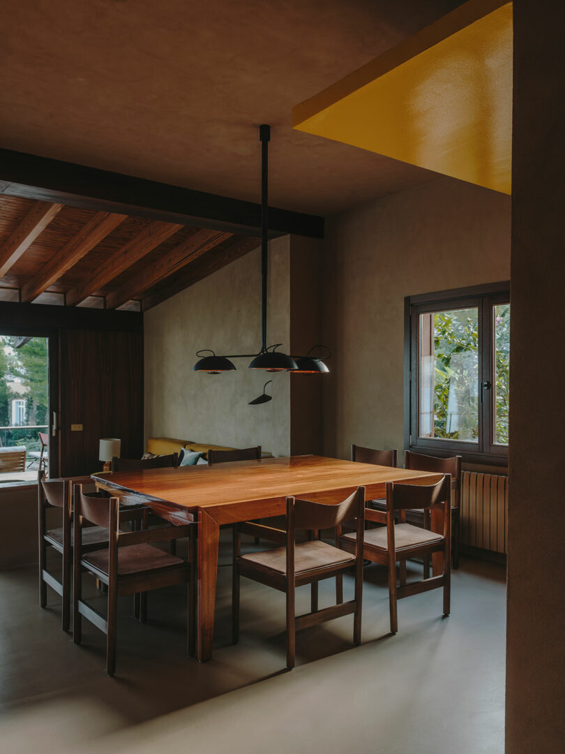A wooden dining table with six matching chairs sits under a modern black pendant light in a room with wooden beams, beige walls, and a window showing greenery outside.