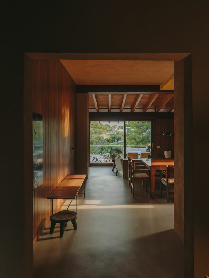 View through a doorway into a wooden dining room with a table, chairs, bench, and natural light coming from large windows overlooking greenery outside.