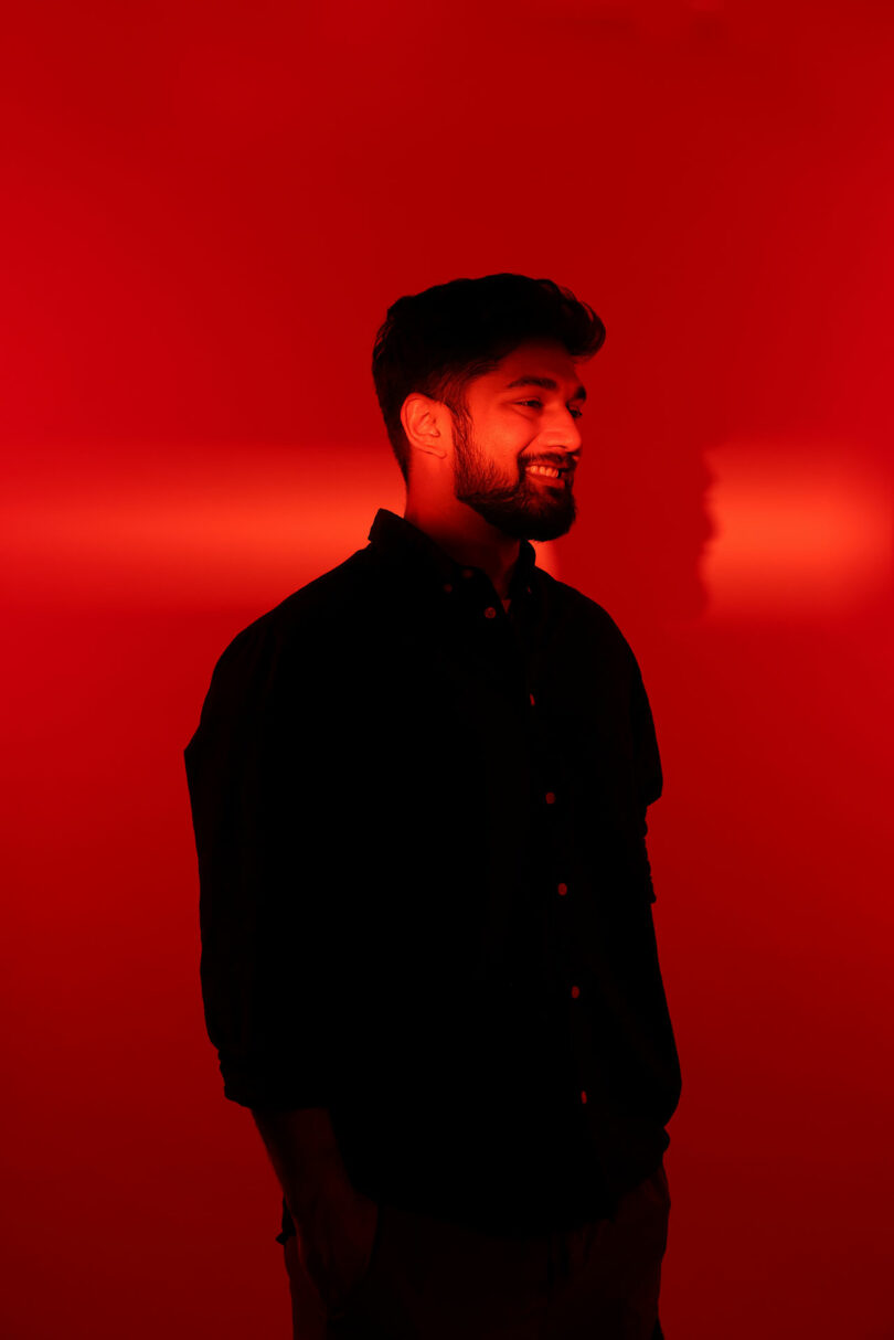 A man with a beard and mustache stands smiling in front of a red background, wearing a dark button-up shirt.