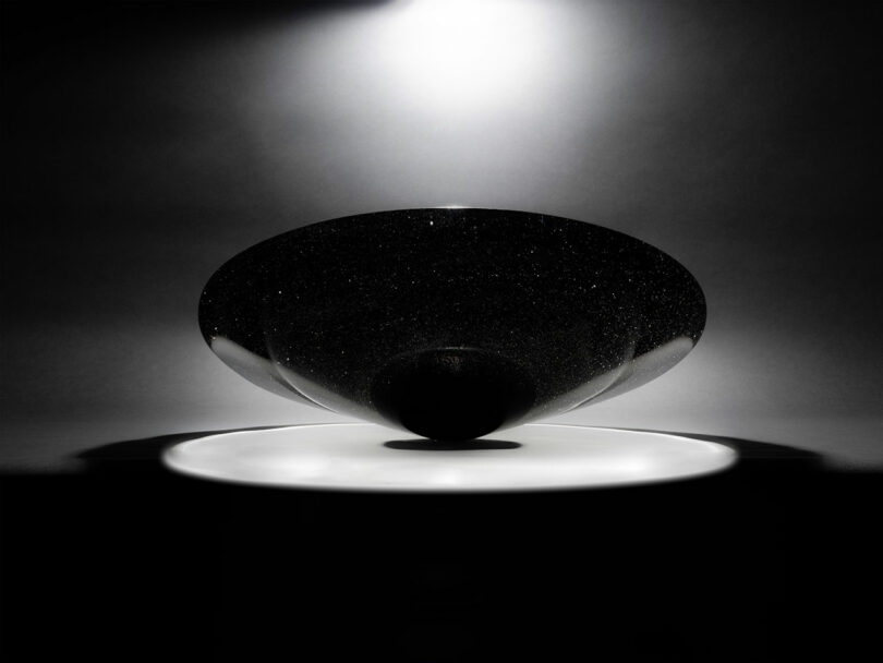 A black, glossy, bowl-shaped object—reminiscent of Claesson Koivisto Rune’s minimalist designs—rests on a white circular surface, illuminated overhead against a dark background.