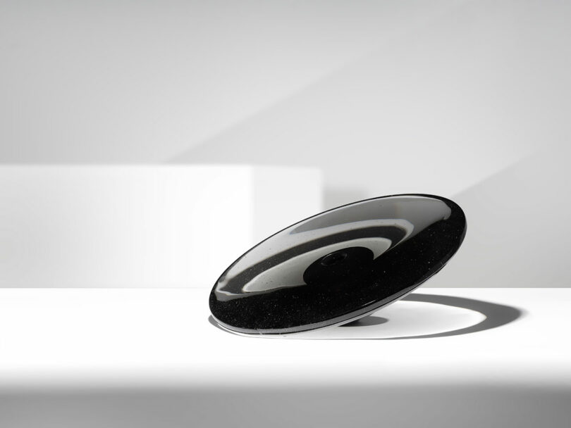 A black and white, disk-shaped object with a reflective surface, reminiscent of Claesson Koivisto Rune's minimalist designs, rests on a white surface in a minimalist setting.
