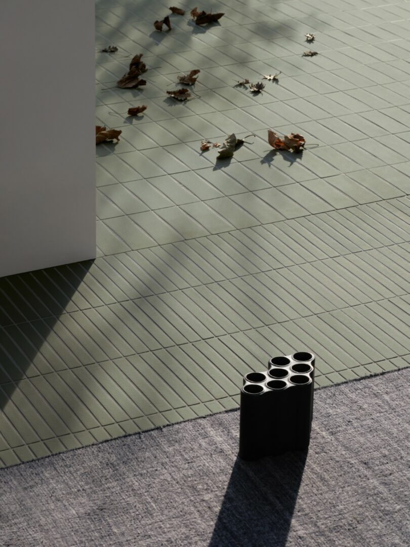 A black, multi-chambered vase sits on a gray rug next to green tiled flooring with scattered dry leaves and sunlight casting shadows