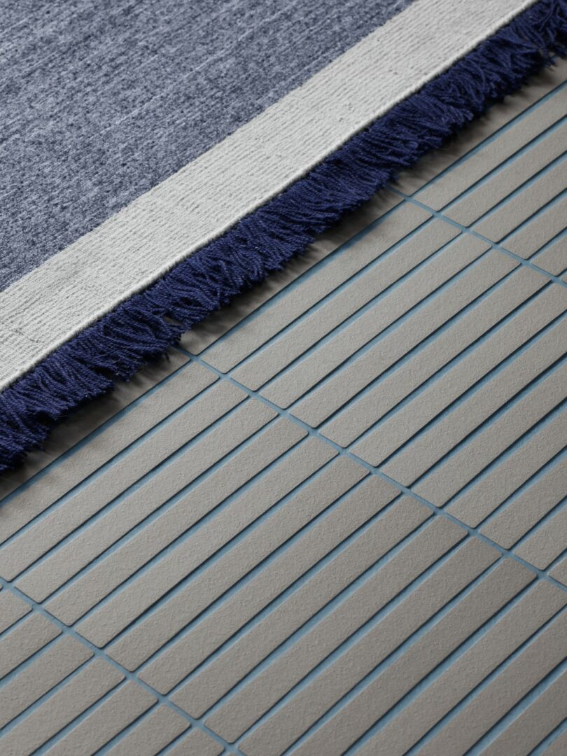 A close-up of a blue and white fringed rug partially covering a gray tiled floor with thin blue grout lines