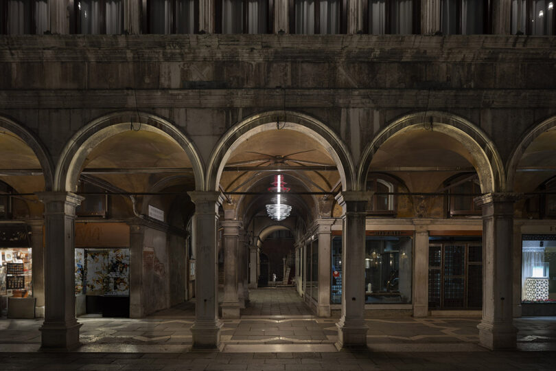 A stone arcade with three illuminated arches at night, flanked by shop windows and lit by overhead lights, with a chandelier and neon sign visible in the passageway.