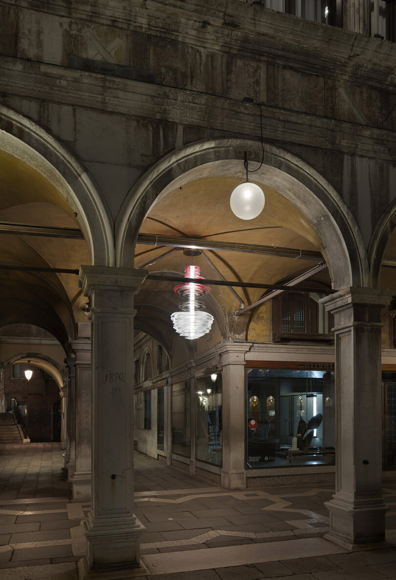 Stone archways at night, illuminated by spherical and spiral hanging lights, with shop windows and a tiled walkway beneath.