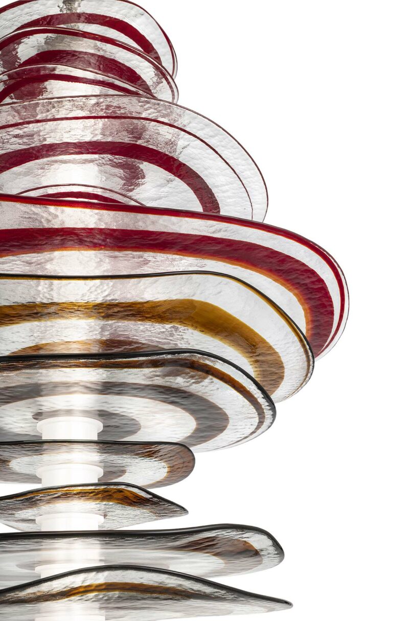 Stacked, circular glass discs in shades of red, amber, and clear tones are arranged in an abstract, sculptural form against a white background.