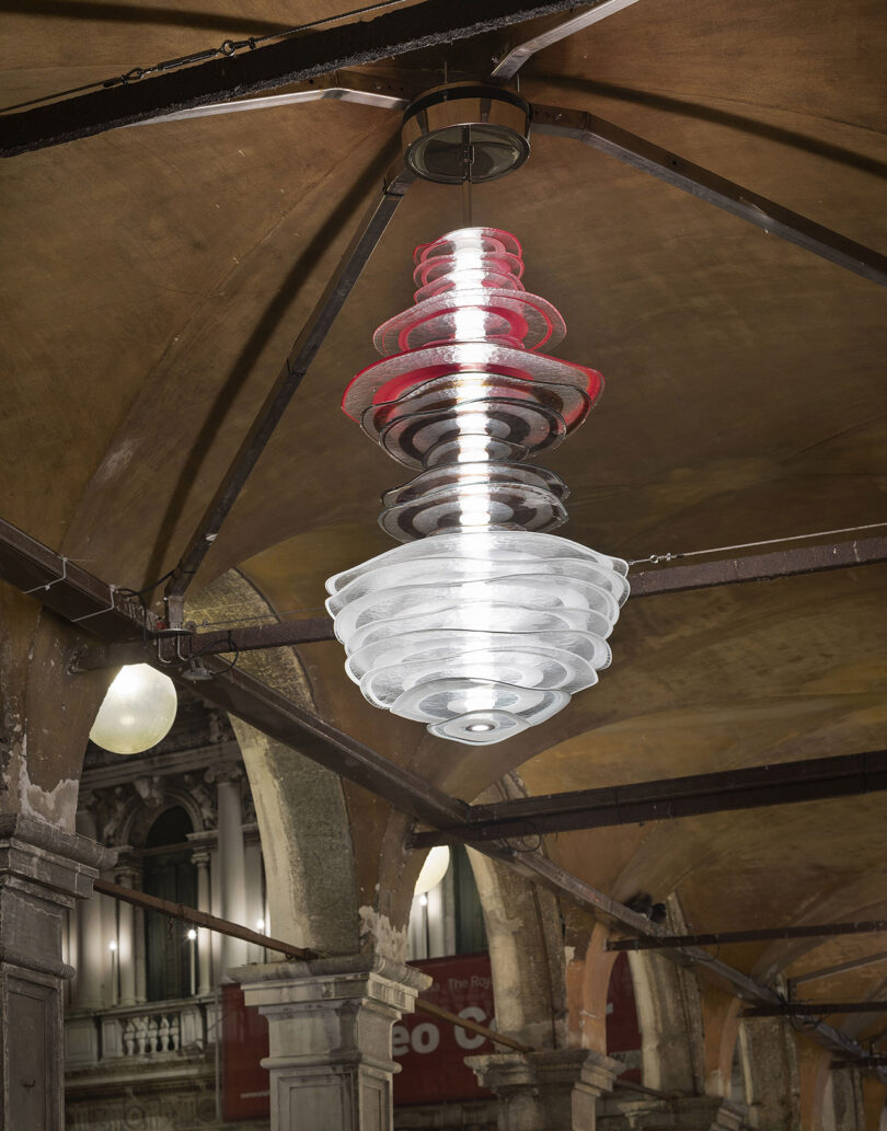 A modern, layered pendant light fixture with red and white illumination hangs from a vaulted ceiling in an arched, historic architectural setting.
