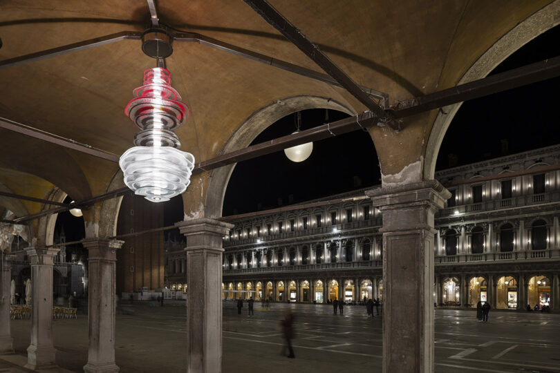 A modern, spiral-shaped light fixture hangs under an arched walkway at night in a historic plaza with illuminated buildings in the background.