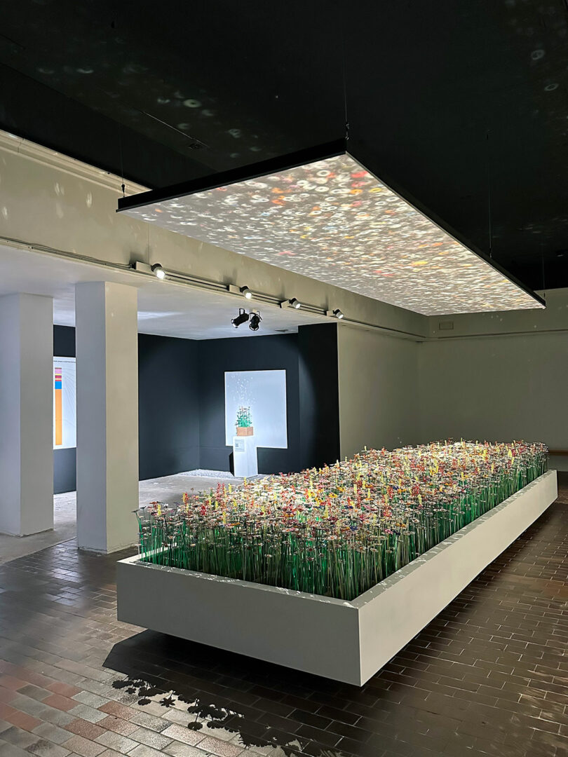A modern art installation features a rectangular bed of colorful artificial flowers, with a large illuminated panel suspended above in a gallery space, beautifully reflecting diversiveness and mo man tai's spirit.
