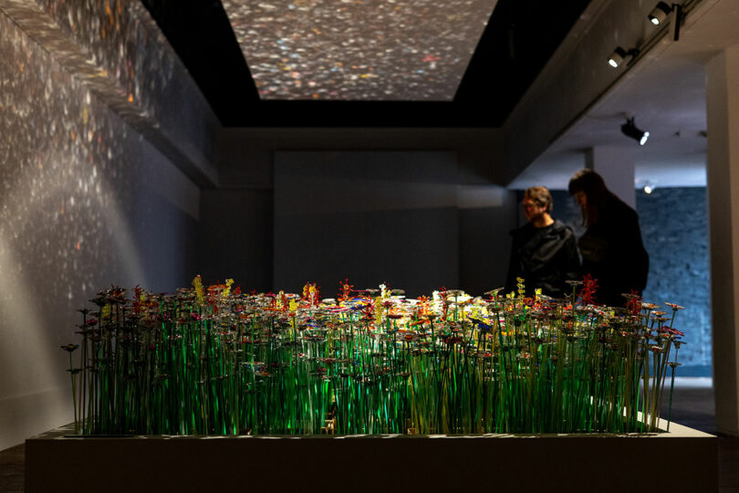 Two people observe an art installation featuring a dense arrangement of colorful artificial flowers, with light projections illuminating the walls and ceiling, beautifully reflecting diversiveness mo man tai.