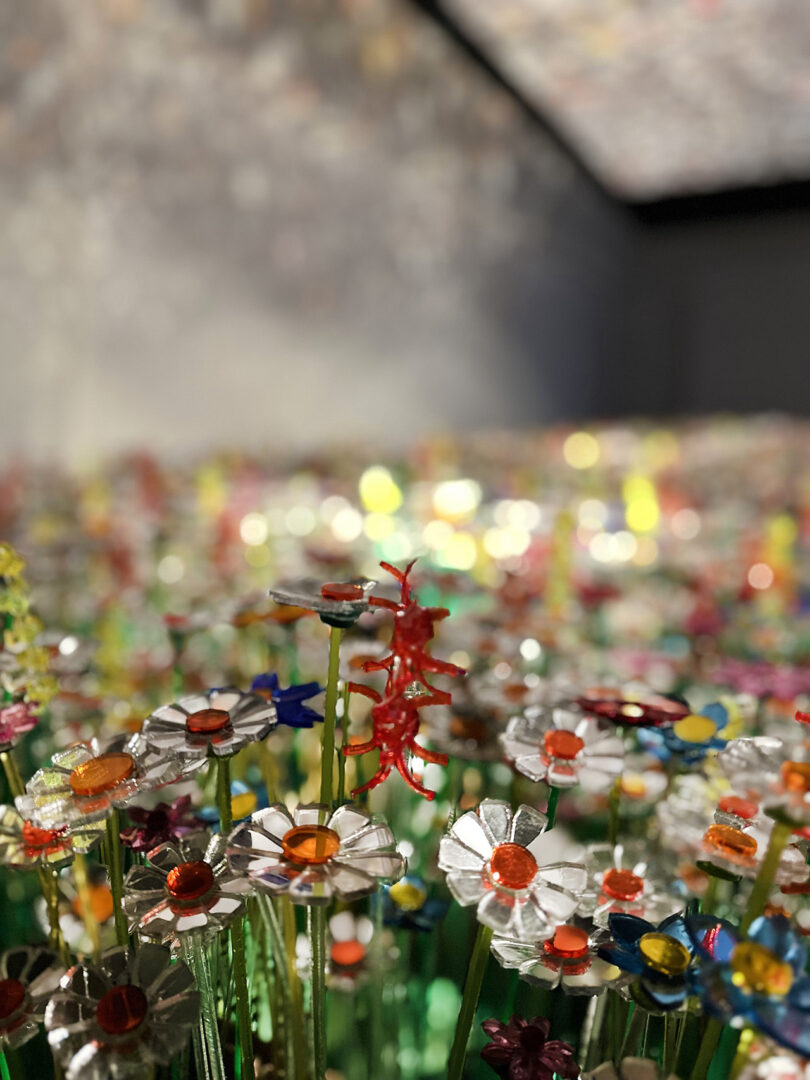 A field of colorful, artificial flowers made from shiny materials is shown indoors, with a blurred background and light streaming in from above.