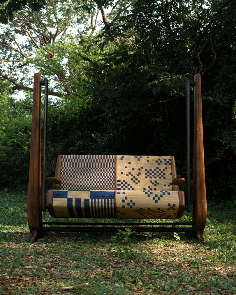 A wooden swing bench with a woven seat and backrest featuring different patterns, set outdoors on grass with trees in the background.