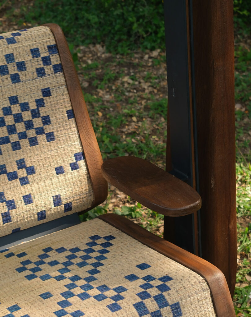 A close-up of a woven chair with a blue geometric pattern, wooden frame, and a small wooden armrest, outdoors on grass.