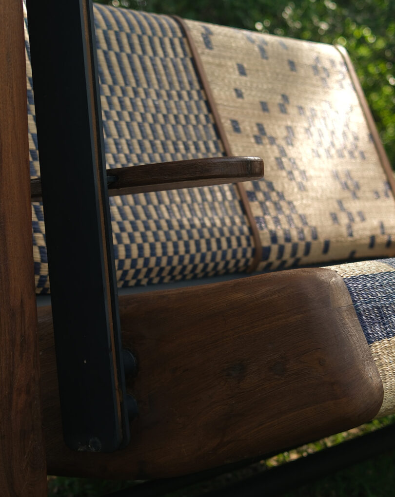 Close-up of a wooden outdoor bench with woven blue and beige seat and backrest, photographed in sunlight with a background of green grass.